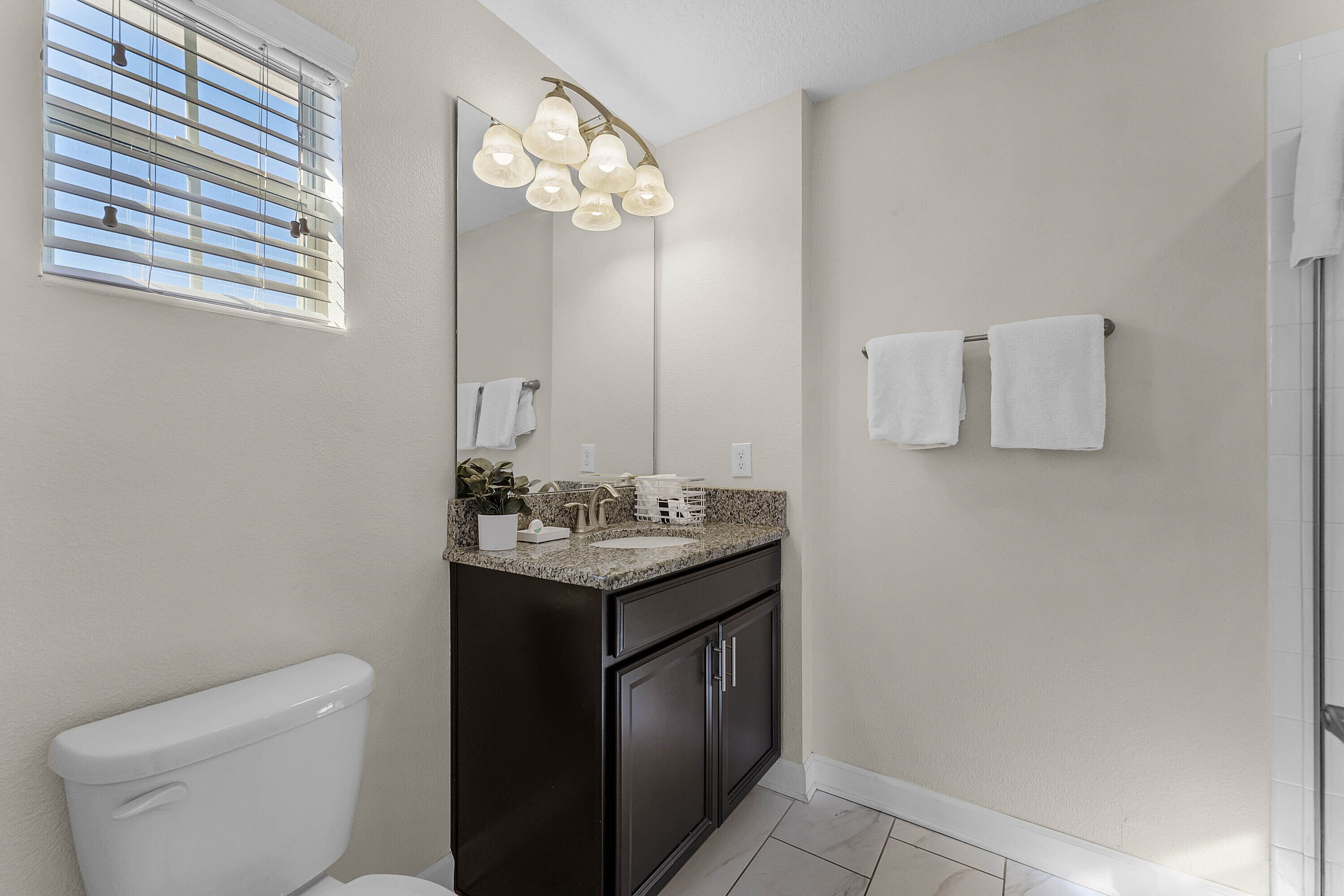 Un baño con inodoro, lavamanos, mueble oscuro, espejo y dos toallas en un perchero.