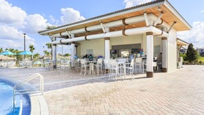 A covered outdoor bar area with tables and chairs, a pool, and a clear sky.
