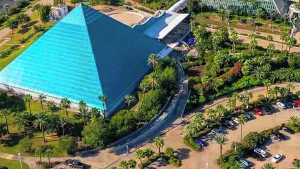 Un grand bâtiment en forme de pyramide, surmonté d’un toit bleu, entouré de verdure et bordé par une étendue d’eau.