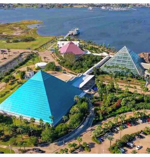 Un gran edificio en forma de pirámide con techo azul, rodeado de vegetación y una masa de agua.