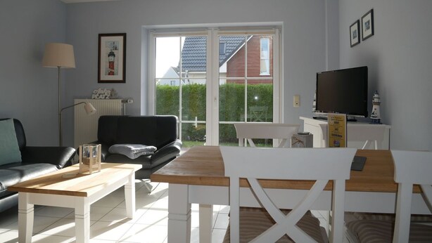A living room with a sofa, coffee table, and TV stand. There is a window with a view of a house and a hedge.