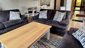 Una sala moderna con dos sofás oscuros, una mesa de centro de madera y cojines estampados. Se ve, al fondo, un comedor con sillas blancas y una mesa.