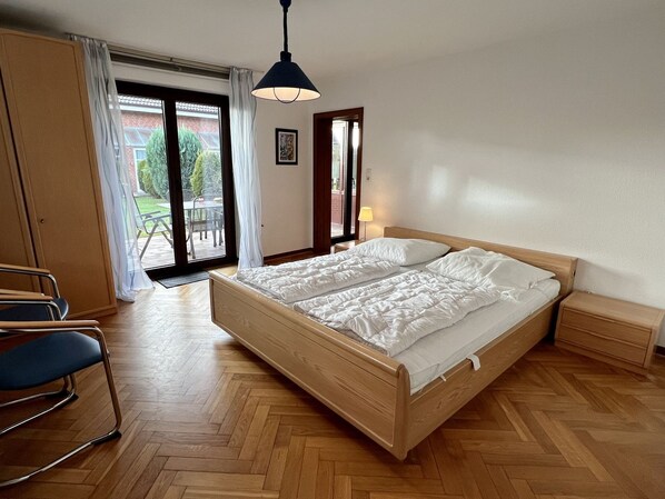 A bedroom with a wooden bed, a chair, a nightstand, a lamp, and a view of the outdoors through a glass door.