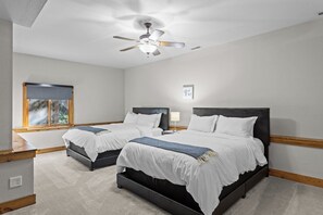 Une chambre avec deux lits, un ventilateur de plafond, une fenêtre avec des stores et un petit tableau accroché au mur.