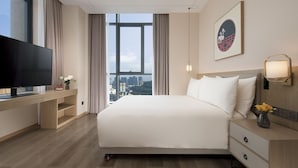 Habitación de hotel con una cama, un televisor sobre un escritorio, vistas de la ciudad y un florero con flores en la mesita de noche.
