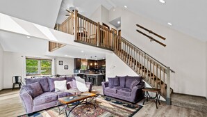 A spacious living area with a high ceiling, a wooden staircase, and a kitchen in the background. - Hot Tub, Large 6 Bed, Game Room, Shuffleboard (Albrightsville)