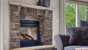 A stone fireplace with a fire, a window with a view of trees, and a gray sofa. - Hot Tub, Large 6 Bed, Game Room, Shuffleboard (Albrightsville)