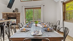 A dining area with a long table set for six, a fireplace, and large windows offering a view of trees. - Hot Tub, Large 6 Bed, Game Room, Shuffleboard (Albrightsville)