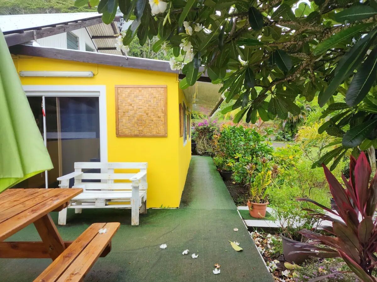 Um edifício amarelo com um banco de madeira e uma mesa, rodeado por plantas em vasos e vegetação.