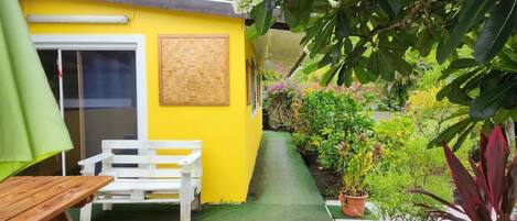 Um edifício amarelo com um banco de madeira e uma mesa, rodeado por plantas em vasos e vegetação.