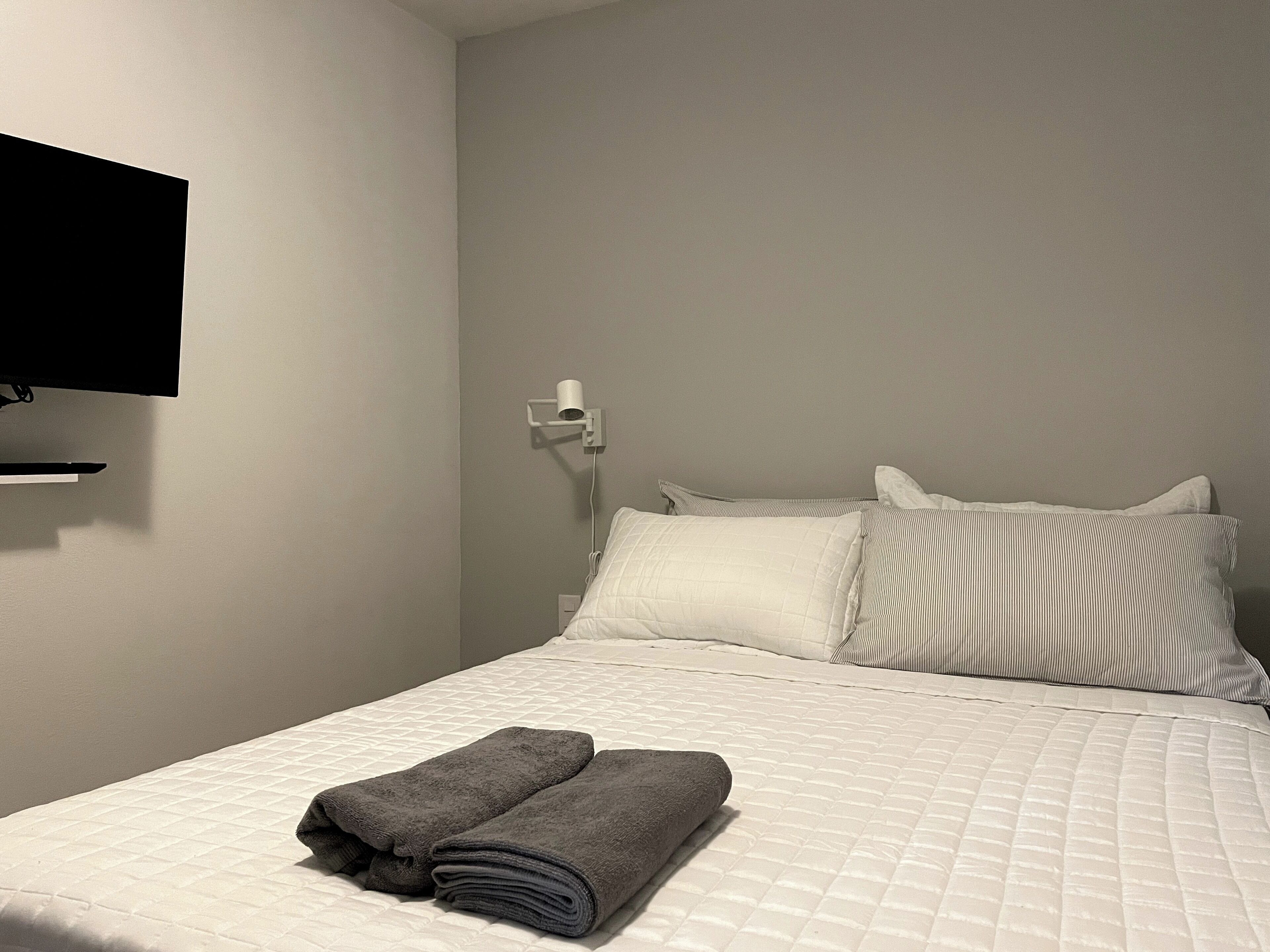 A neatly made bed with white and striped pillows, a rolled-up gray towel, and a white wall-mounted TV.