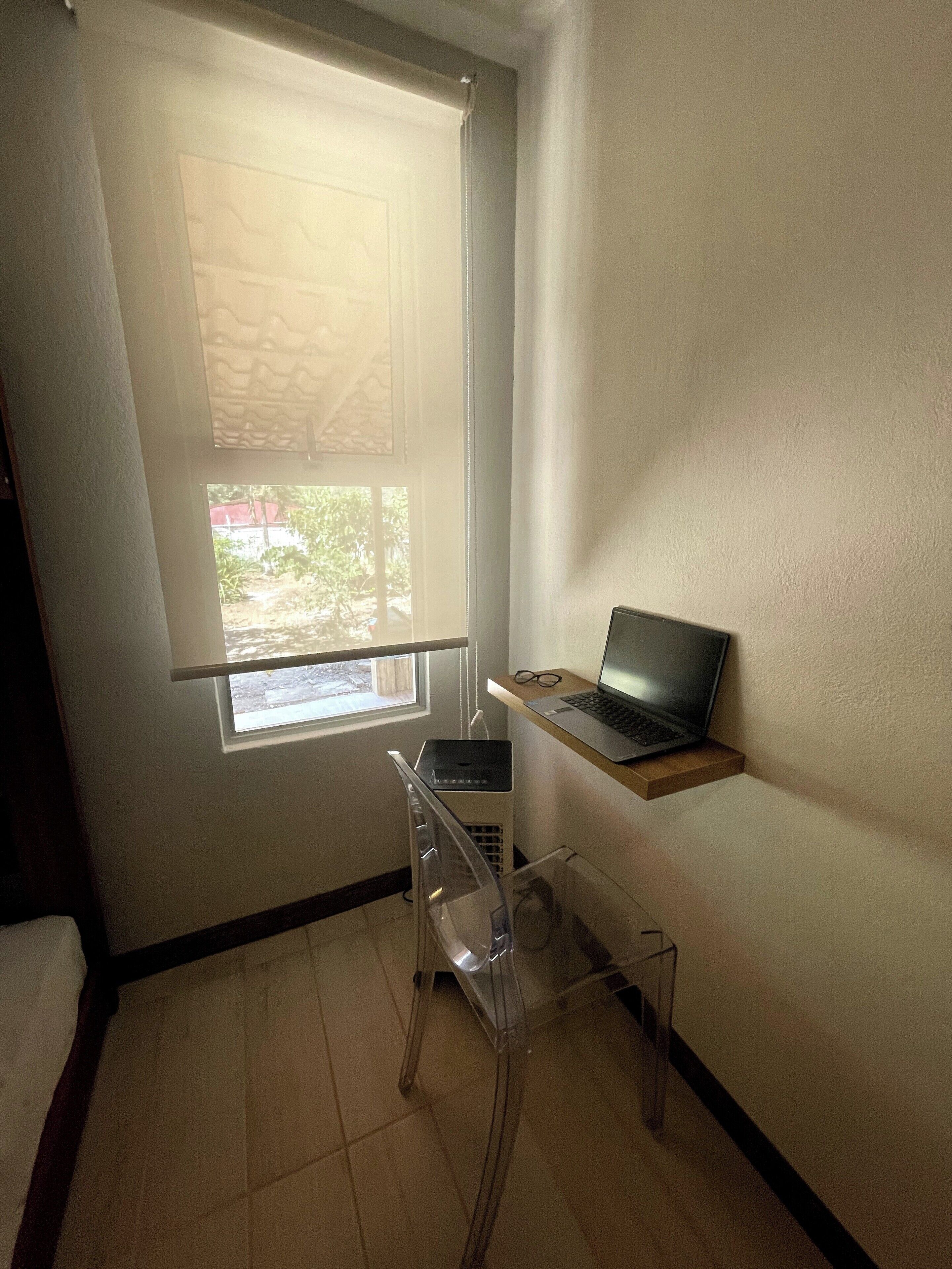 Een kleine kamer met een raam, een laptop op een plank en een transparante stoel.