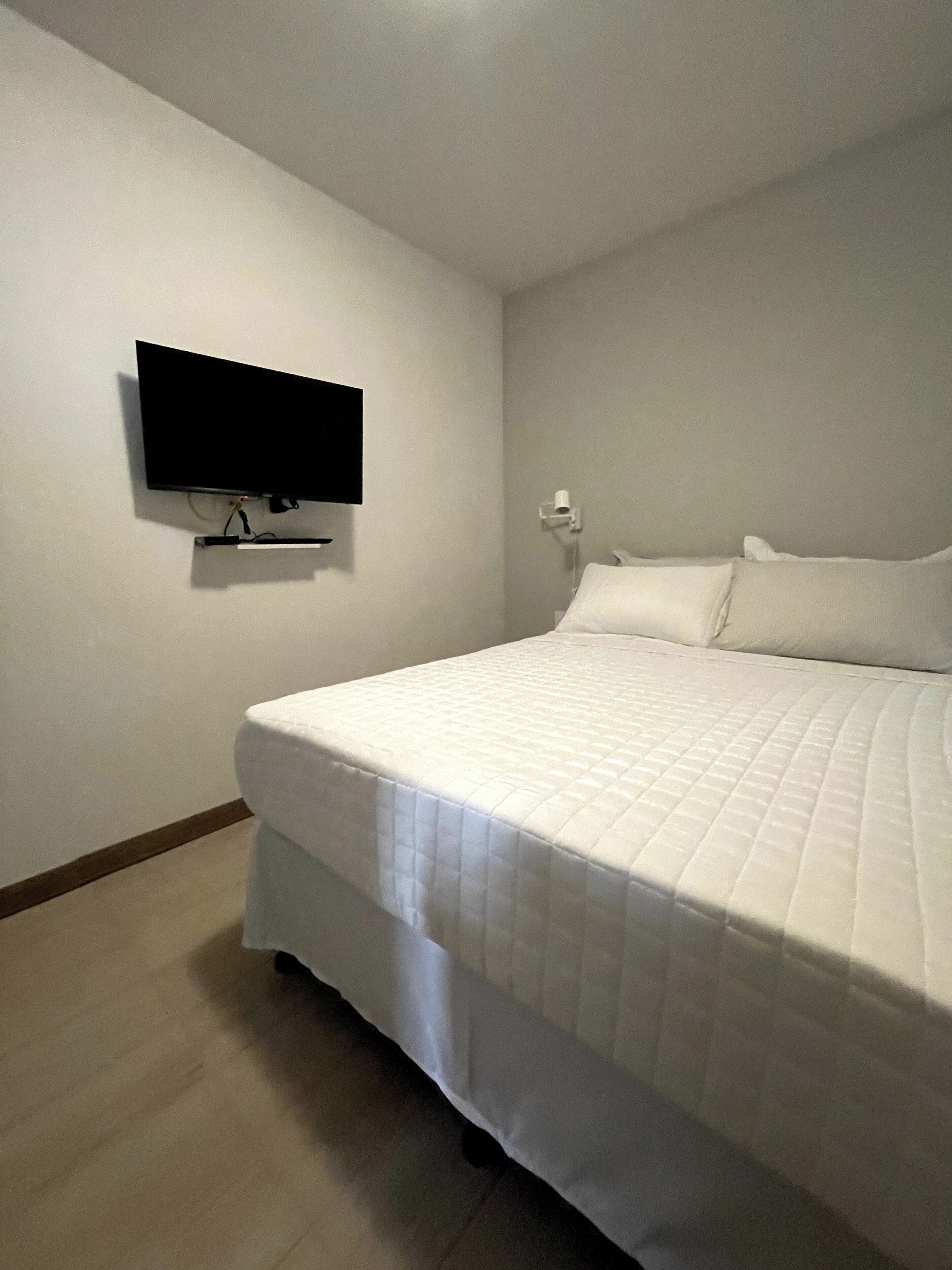 A bedroom with a bed, a flat-screen TV mounted on the wall, and a white wall.