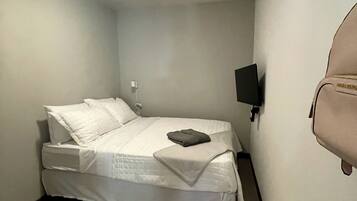 A small, neatly made bed in a corner room with a single wall-mounted TV and a small bag hanging on the wall.