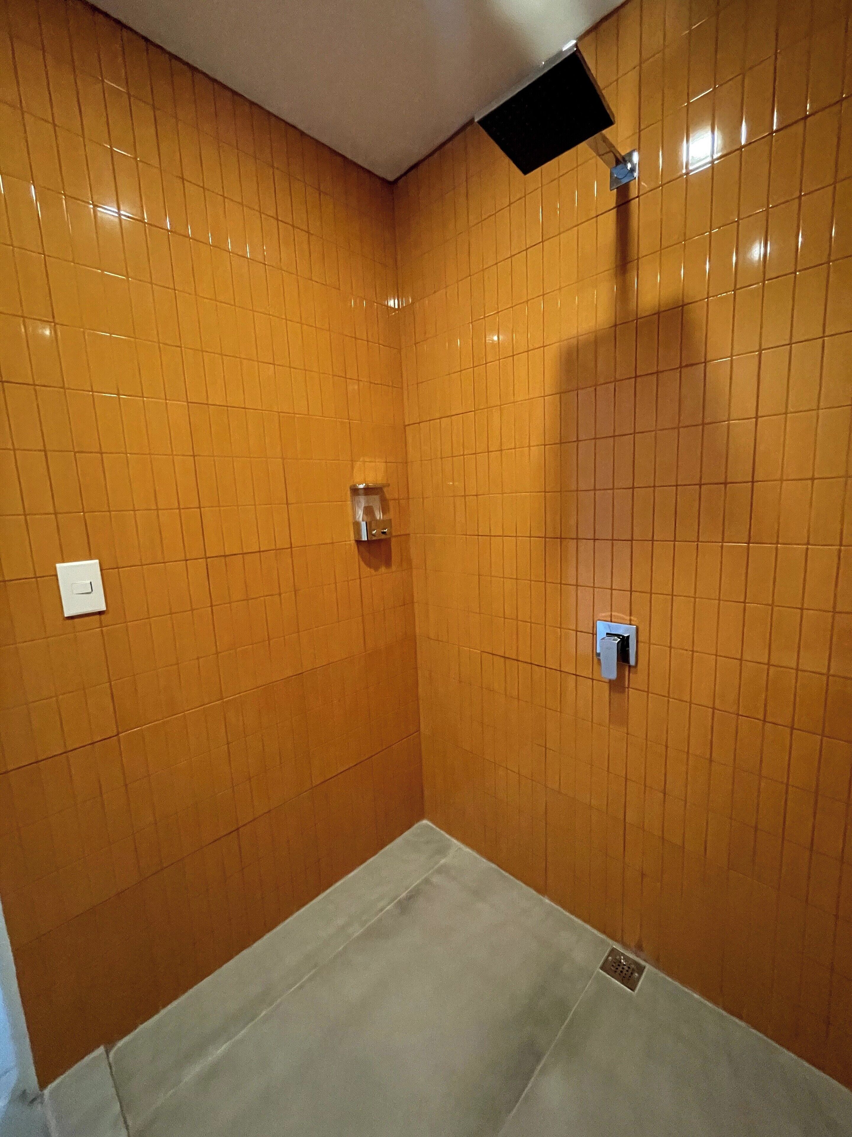 A small, tiled shower room with a single showerhead and a light switch on the wall.