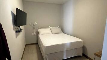 Une petite chambre propre, avec un lit simple, un téléviseur à écran plat fixé au mur et une petite table avec un verre.