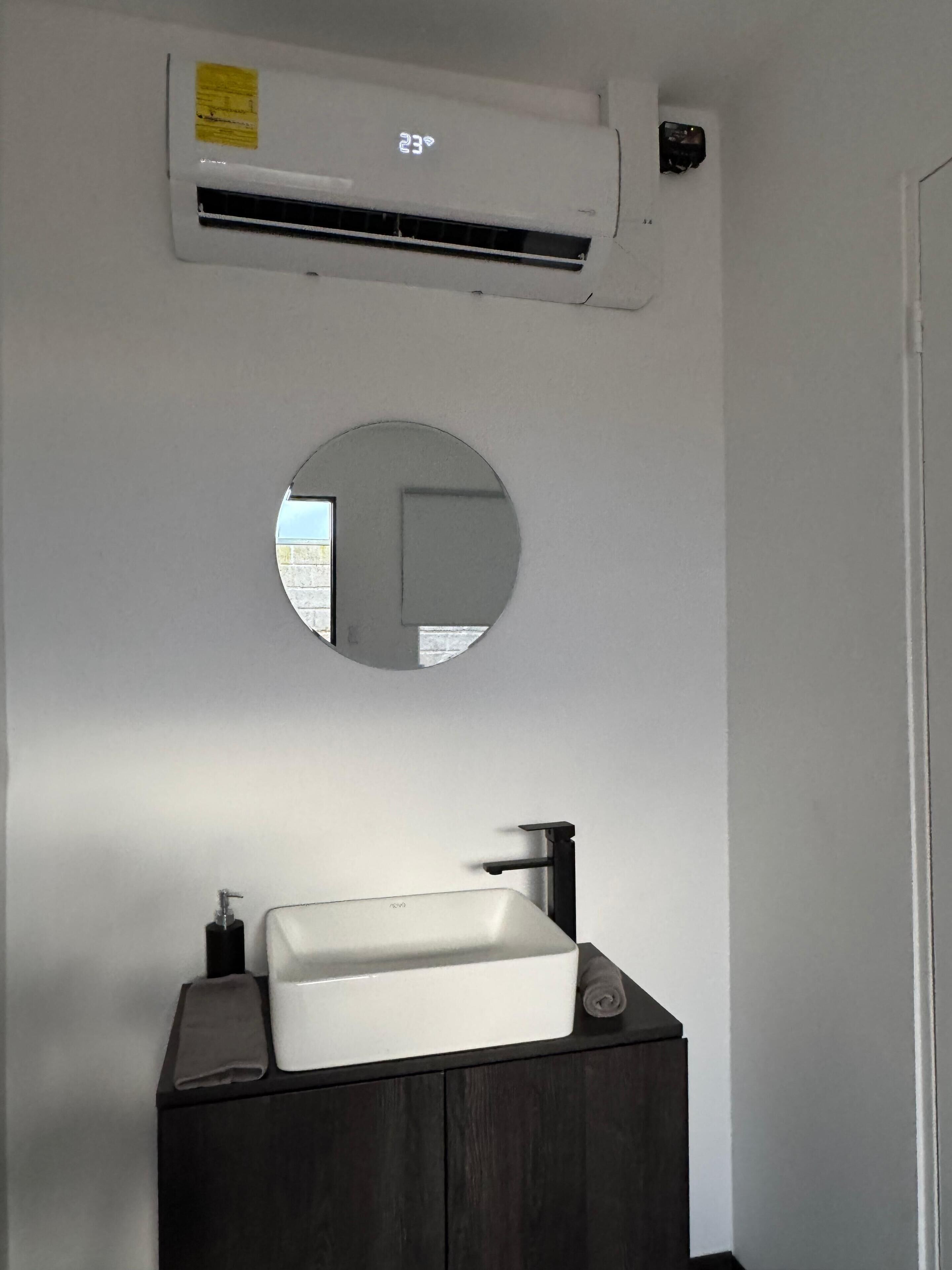 A bathroom with a white sink, dark wooden vanity, a round mirror, and an air conditioning unit above the mirror.