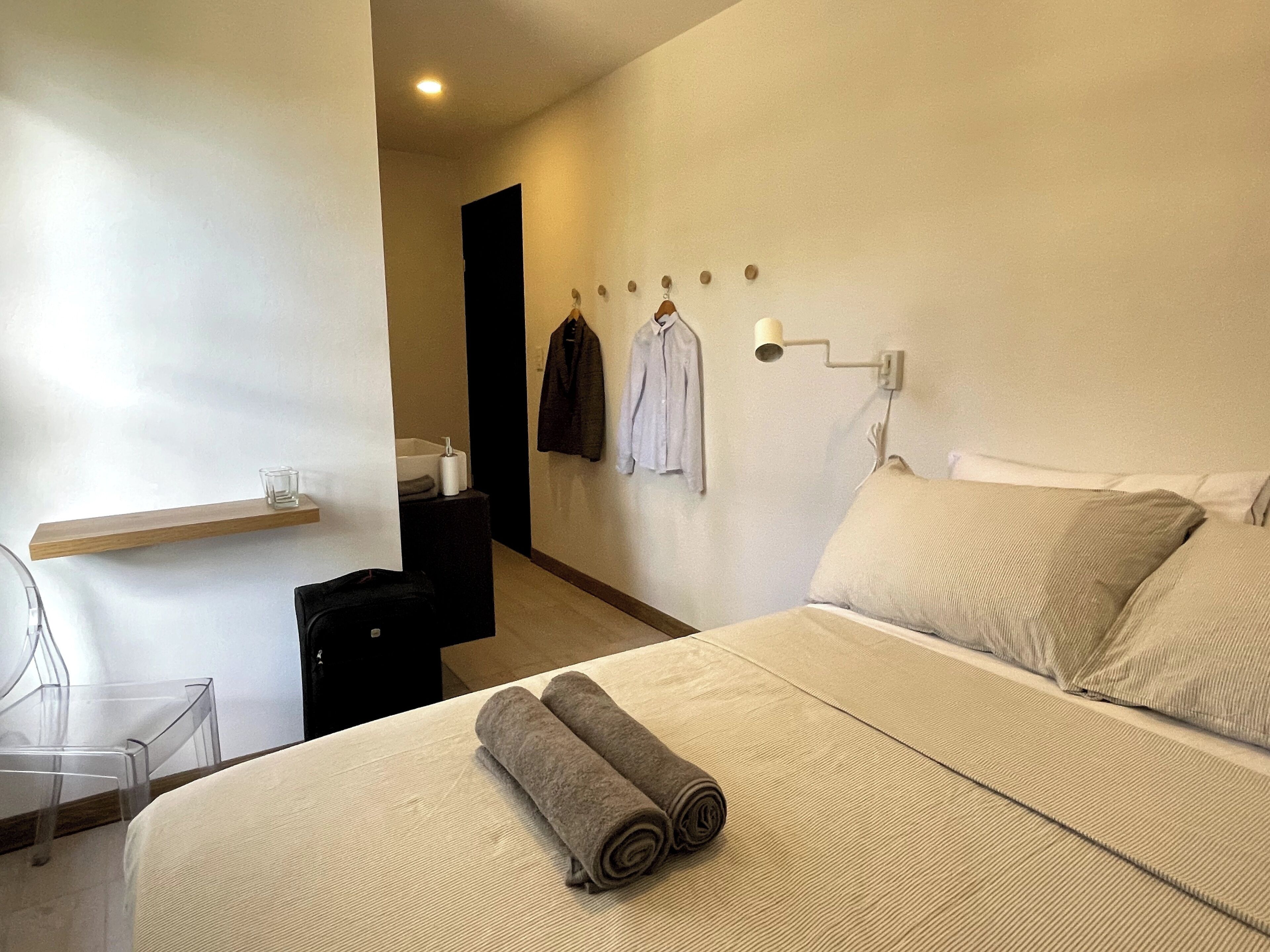 A hotel room with a bed, a chair, a small table, a shelf, and a wall-mounted towel rack.