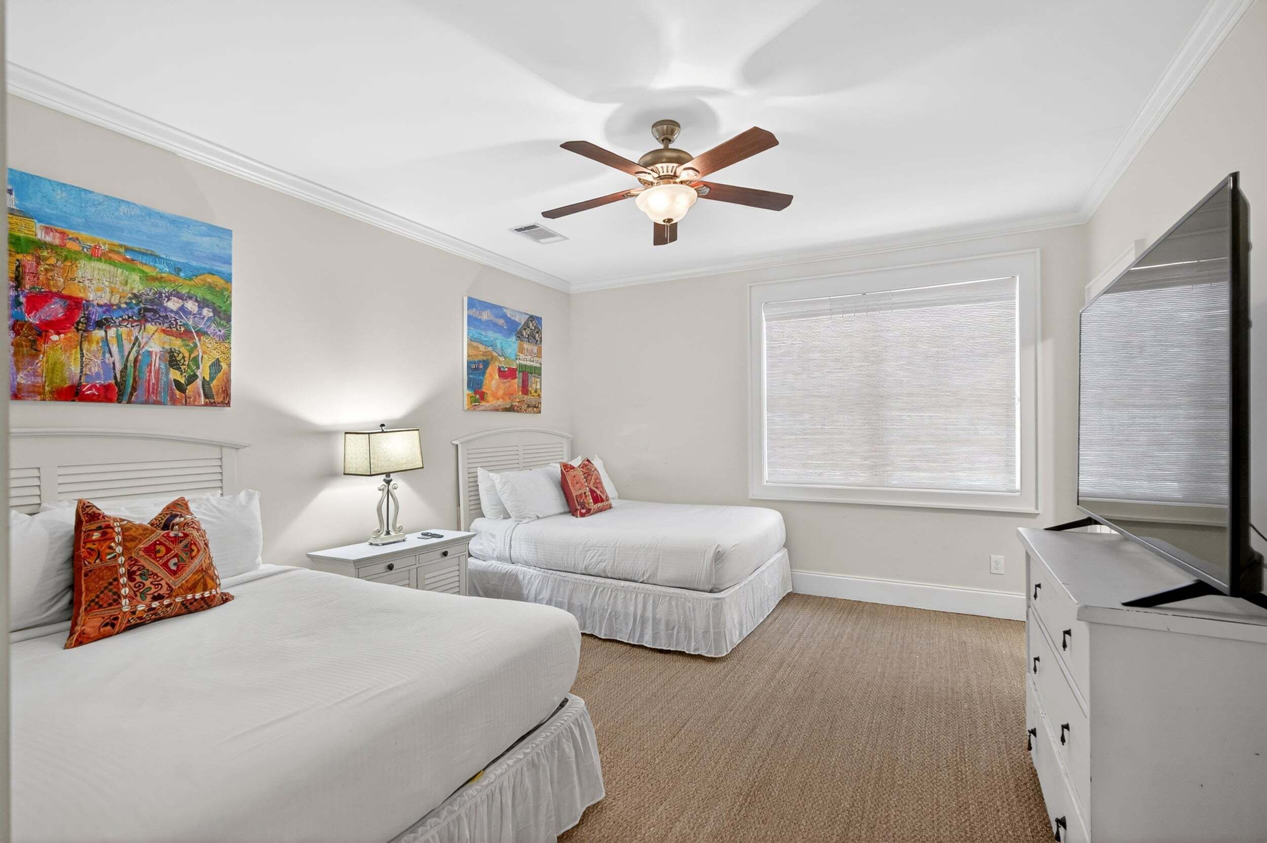 A bedroom with two beds, a ceiling fan, a TV, and artwork on the walls.