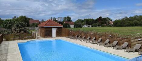 A rectangular swimming pool with a wooden deck and lounge chairs, surrounded by a wooden fence and a small building with a red roof.