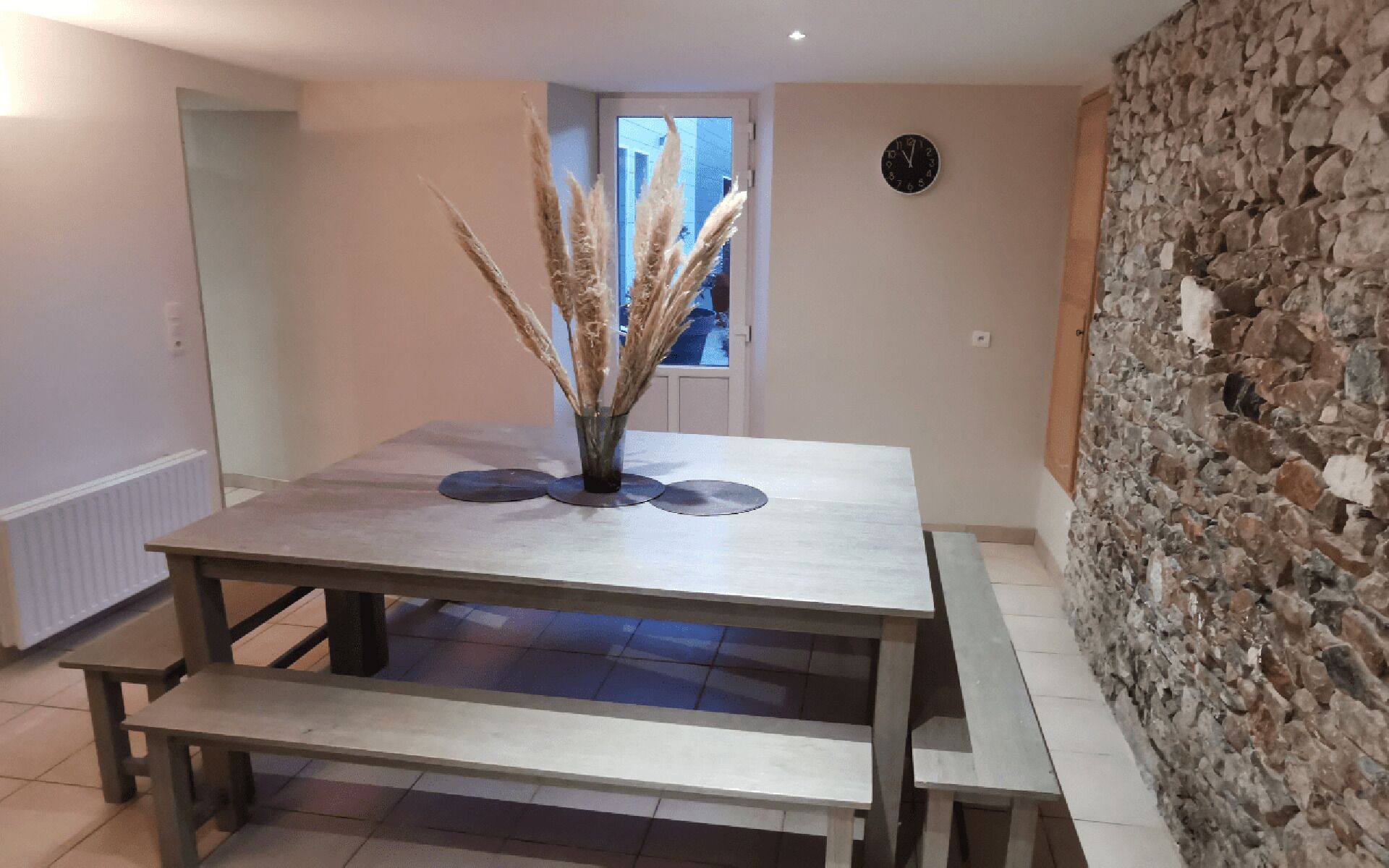 A dining area with a wooden table and benches, a vase with dried plants, and a stone wall.