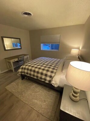 Relax in a serene space with a comfortable bed, bedside table, and desk, all set against a neutral backdrop.