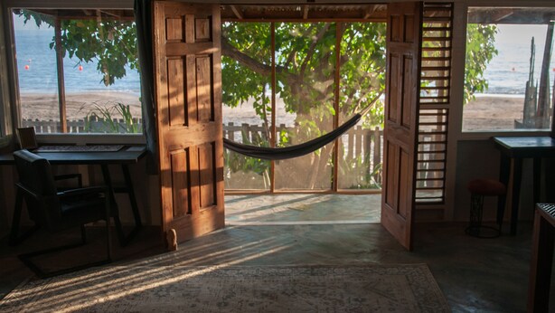 A room with a hammock, wooden doors, and a view of the beach.