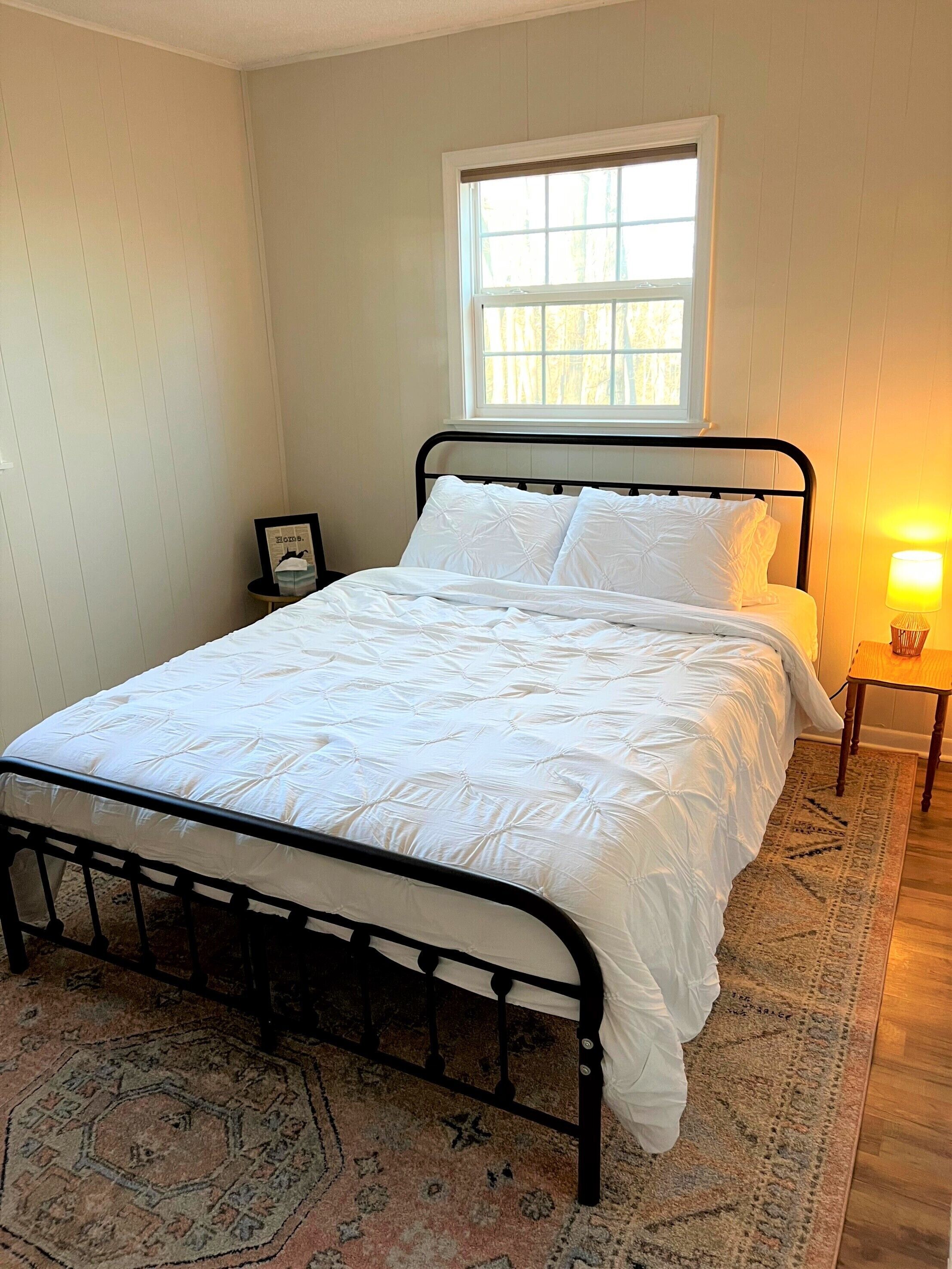 A neatly made bed with a white duvet and pillows, a small bedside table with a lamp, and a window with a view of trees.