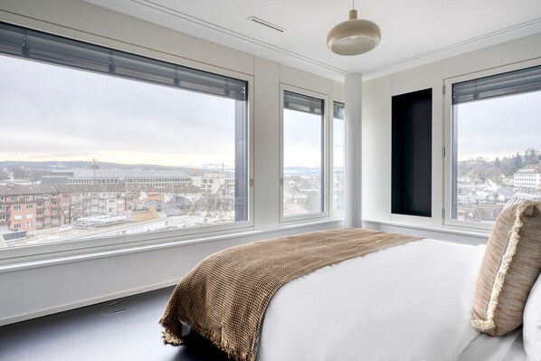 Un dormitorio moderno con ventanas amplias, una cama con manta texturizada y vista a un paisaje urbano.