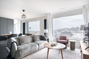 Una sala de estar moderna con un sofá gris, una mesa redonda de centro y un sillón rosa. Amplias ventanas ofrecen vistas a un paisaje urbano.