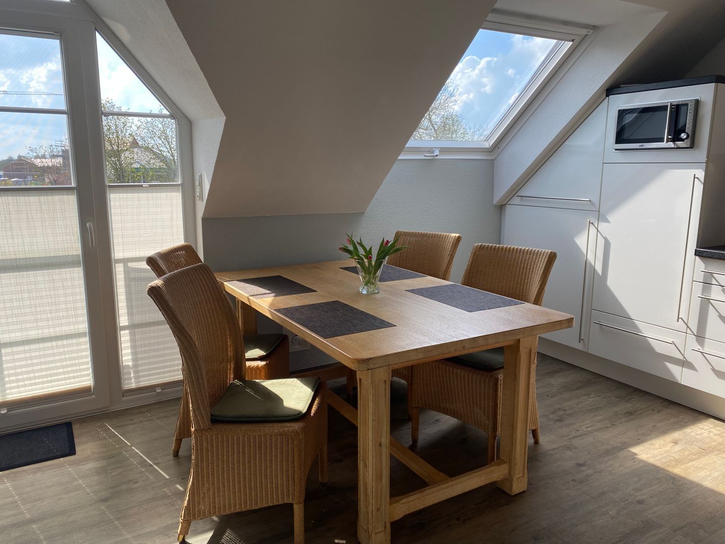 Ein Essbereich mit Holztisch und Korbstühlen, einer kleinen Vase mit Blumen sowie eine Küche mit weißen Schränken und einer Mikrowelle.