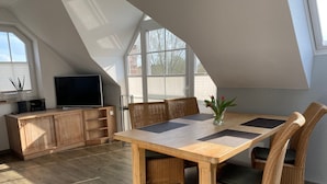 Un comedor con mesa de madera y sillas de mimbre, un televisor sobre un mueble de madera, y ventanas grandes que dejan entrar luz natural.
