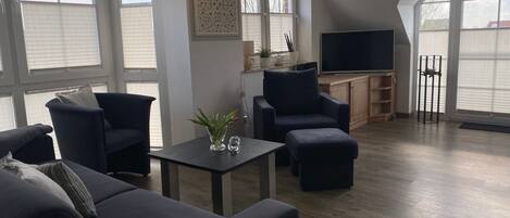 Una sala de estar moderna con un sofá azul oscuro, un sillón a juego, una mesita con un jarrón de flores y un televisor de pantalla plana.
