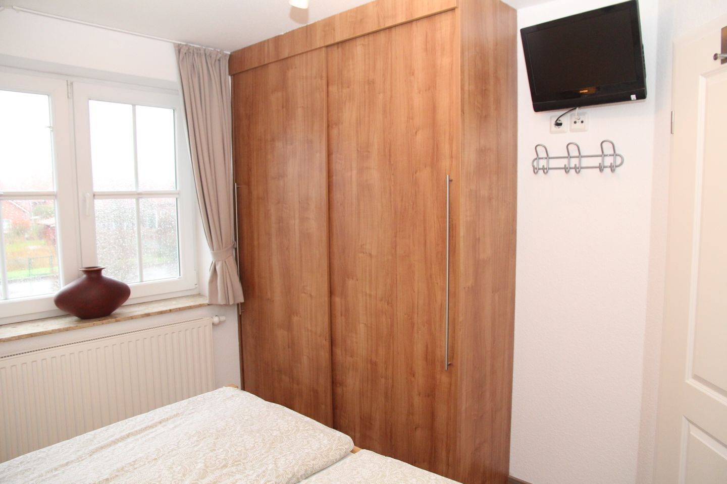 A bedroom with a bed, a wooden wardrobe, a TV mounted on the wall, and a window with curtains.