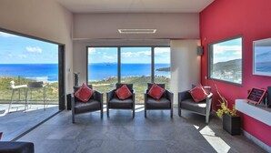 Ett modernt rum med en röd accentvägg, stora fönster med utsikt över havet och en sittgrupp med läderstolar och röda kuddar.