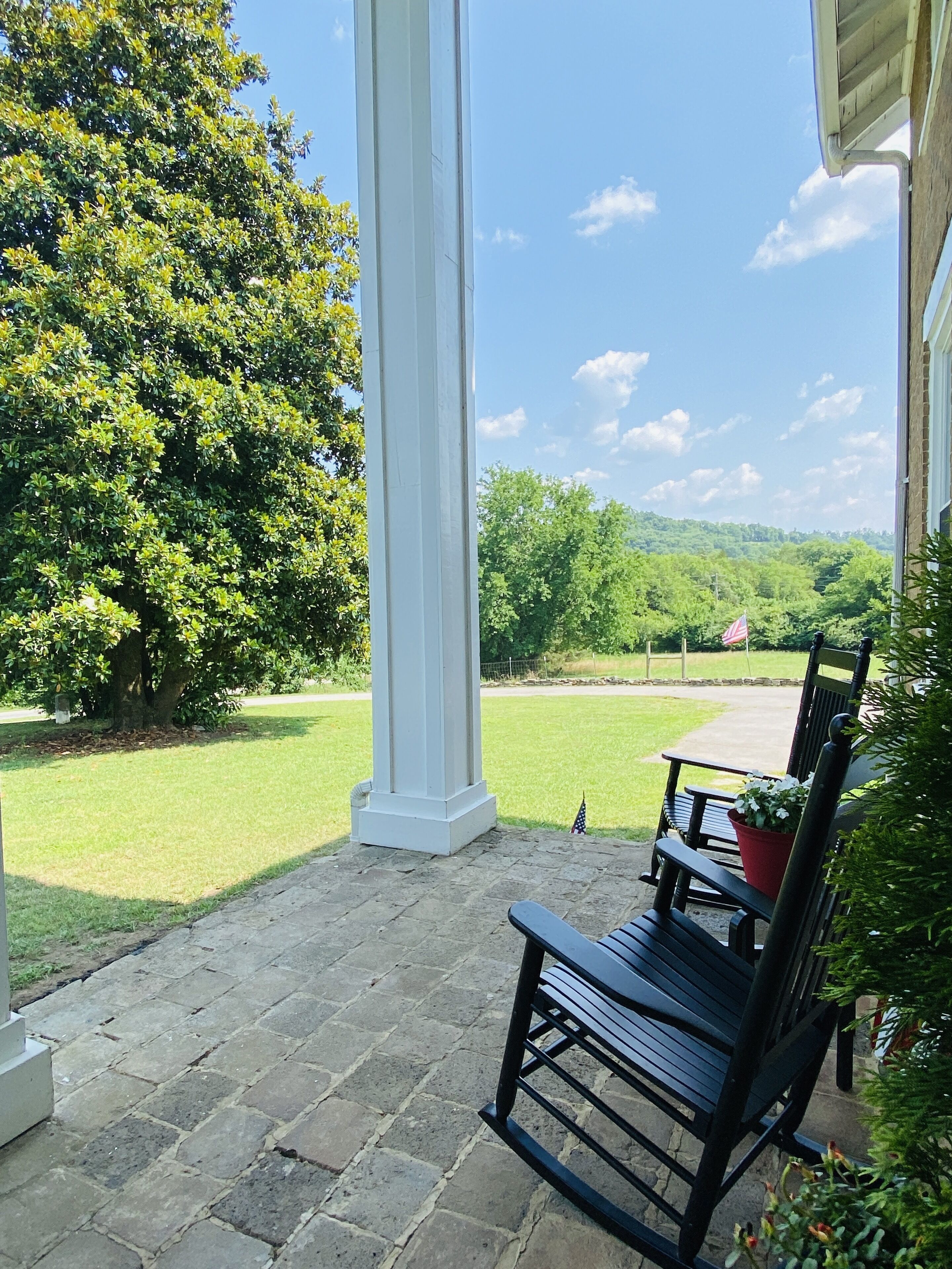 A covered patio area with a stone floor, a wooden bench, and a view of a grassy landscape with trees and a distant hill.
