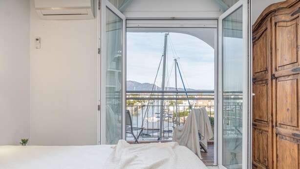 Et soveværelse med en stor seng, et træskab og en skydedør, der fører til en balkon med udsigt over en bro og bjerge.