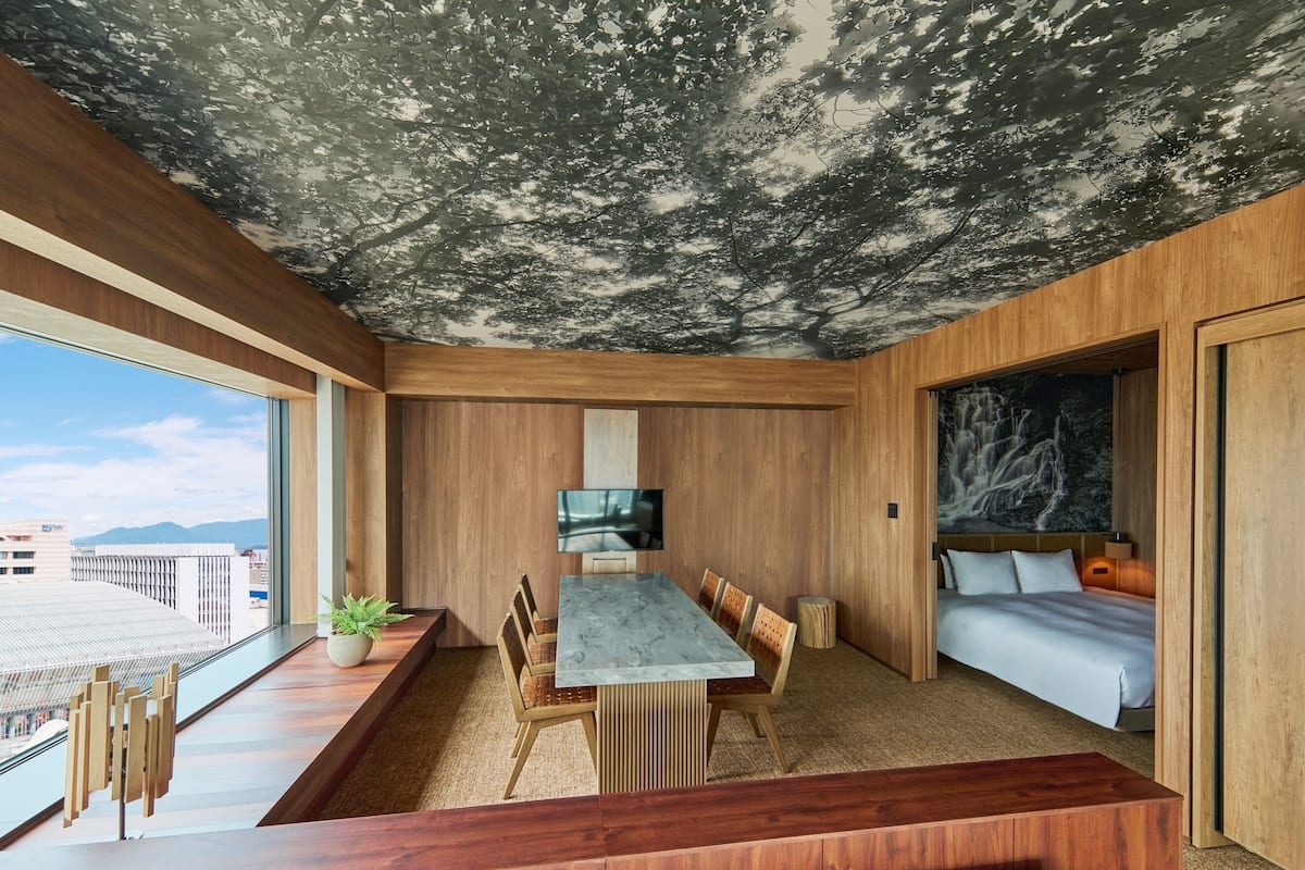 A modern hotel room with a wooden ceiling, a dining area with a marble table, and a large window offering a city view.