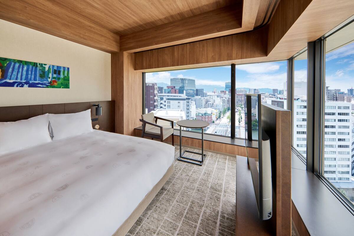 A hotel room with a large bed, a small table, a chair, a TV, and a city view.