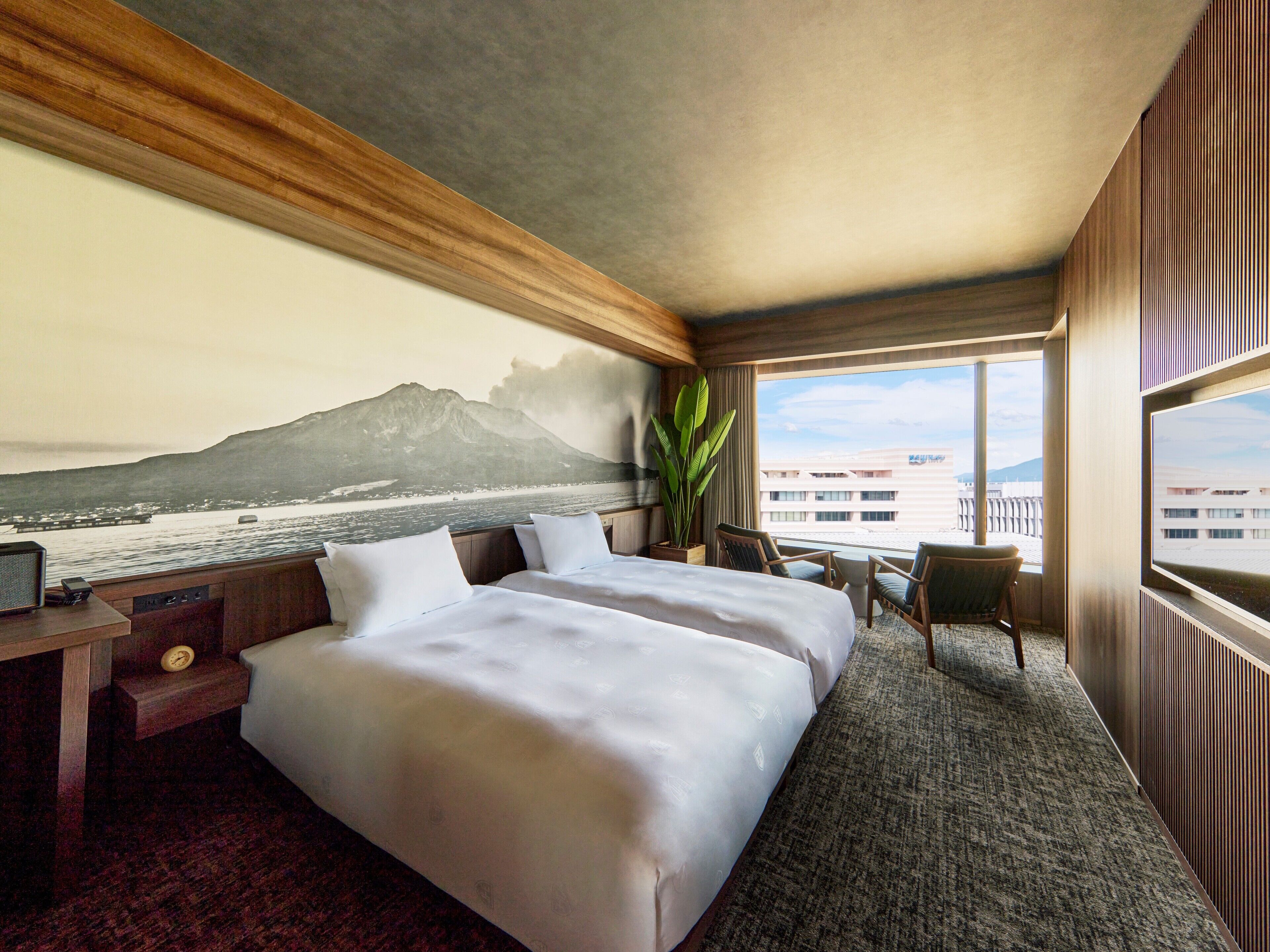 A hotel room with a large bed, a desk with a clock, a chair, a large window with a view of a mountain and water, and a plant.