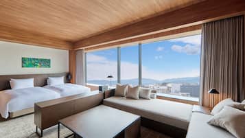 A modern hotel room with a large bed, a sofa, a small table, and a view of the city through large windows.