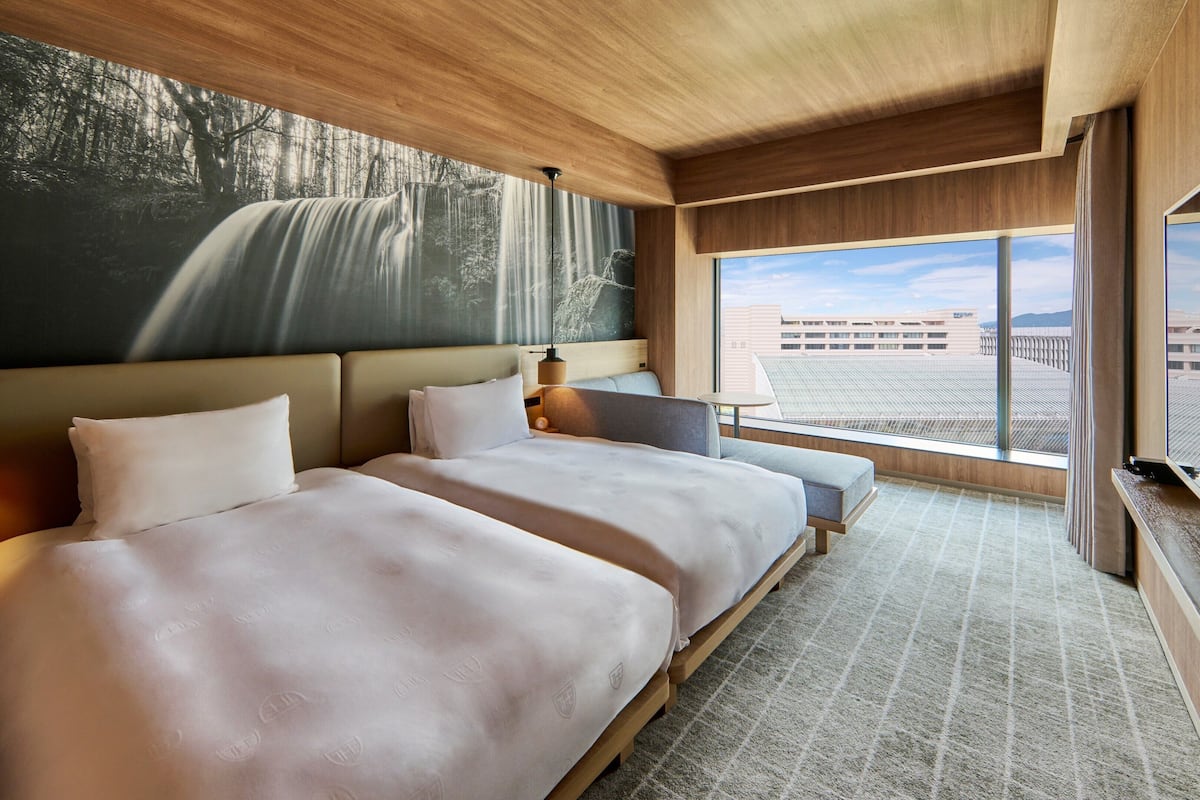 A hotel room with two beds, a large window with a view of a building, and a waterfall painting on the wall.