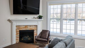 Una sala de estar con chimenea, un televisor montado encima, un sillón cómodo, un sofá y un ventanal con vistas al exterior.