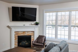 A living room with a fireplace, a television mounted above it, a comfortable armchair, a sofa, and a large window with a view of the outside. - Downtown Sister Bay: 2022 New Construction home (Sister Bay)