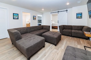 A modern living room with a sectional sofa, ottoman, and a small table. There is a sliding door leading to a bedroom.