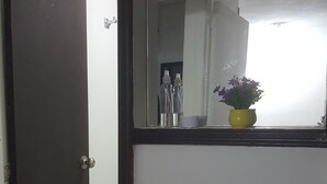 Ein Innenbereich mit einem Flur, einer geschlossenen Tür, einer weißen Wand und einem Fenster mit Blick auf einen Raum, der Flaschen und einen Blumentopf zeigt.