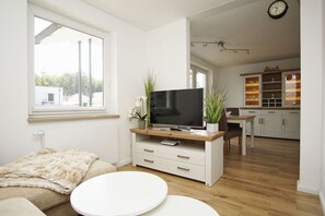 Un salon moderne avec un téléviseur à écran plat posé sur une console en bois, une table basse ronde blanche et un canapé beige. On aperçoit à l’arrière-plan un coin repas avec un meuble de cuisine portant l’inscription « Beach ».