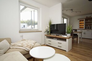 Ett modernt vardagsrum med en soffa, en tv på ett träställ, en matplats med ett bord och stolar samt ett köksområde med skåp och en bänkskiva.