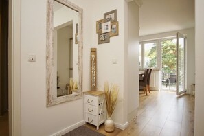 Un couloir avec un grand miroir, un panneau d’accueil, une petite armoire blanche et un vase contenant des plantes séchées.