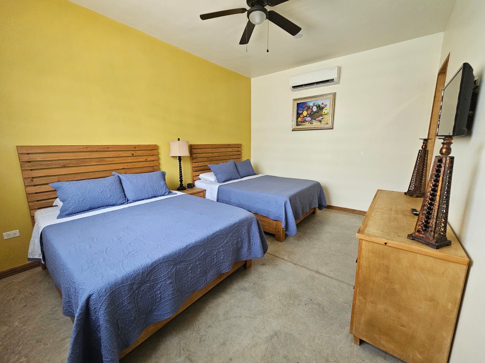 A hotel room with two beds, a ceiling fan, an air conditioning unit, a wooden nightstand, a lamp, a painting, and a wooden dresser with a mirror.